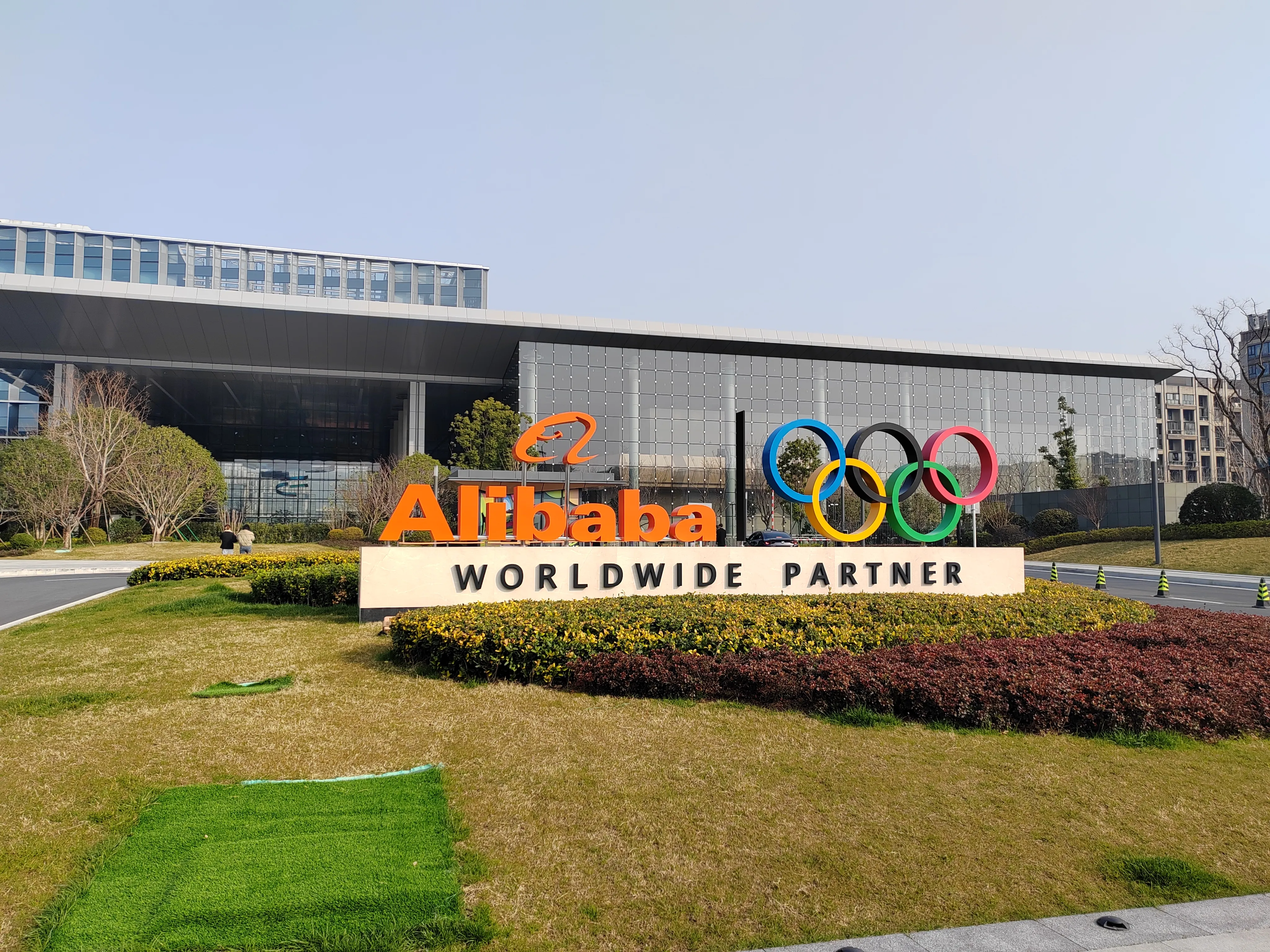 阿里巴巴全球合作伙伴标志，位于一座现代化建筑前。标志上有一个橙色的大‘Alibaba’字样和奥运五环标志。背景是一座玻璃幕墙的建筑，周围有绿色的草坪和灌木丛。天空晴朗，天气良好