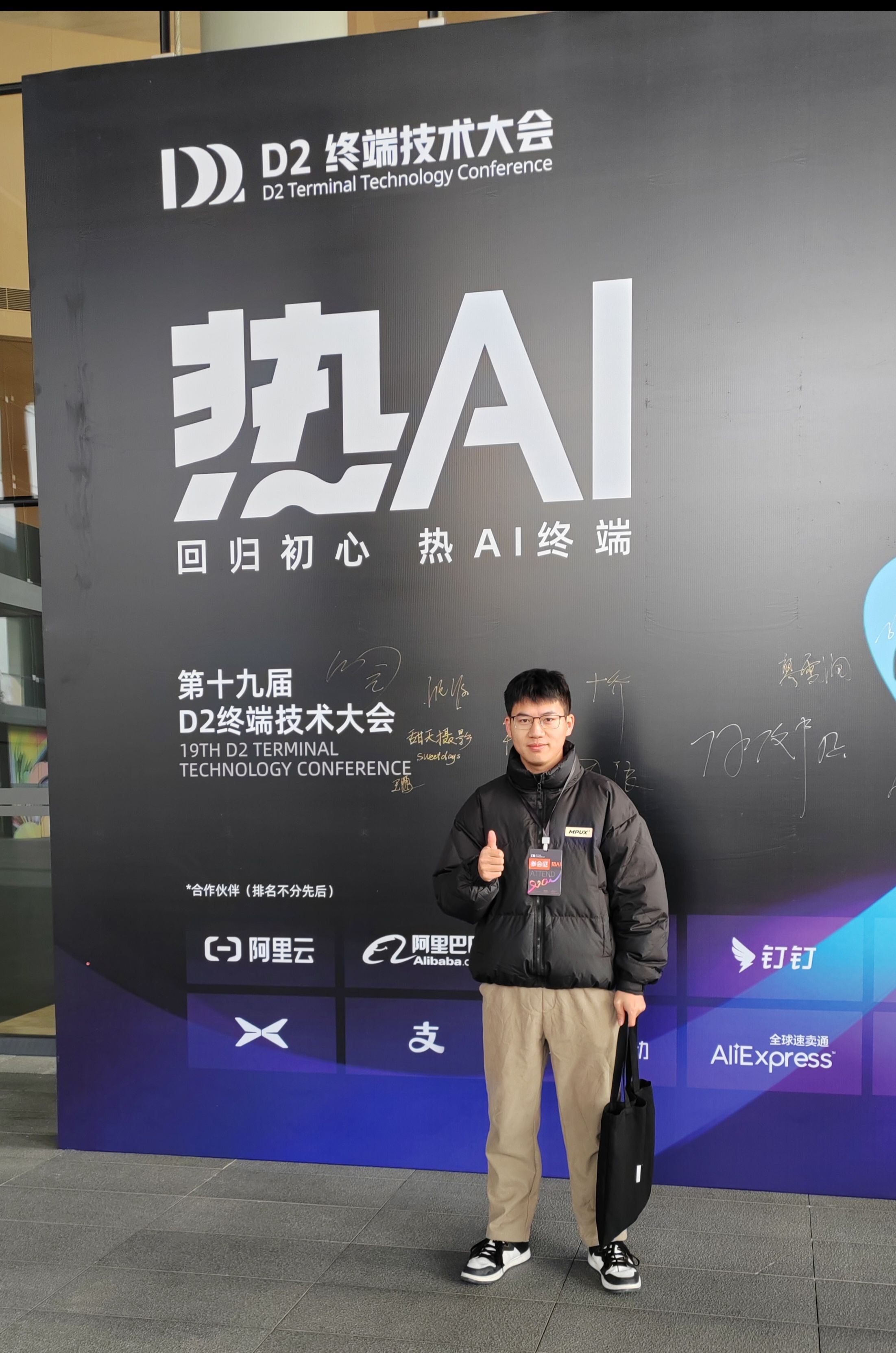 前面站着的是作者本人,黑色外套、卡其色裤子的人,左手提着黑色包,站在 “第十九届 D2 终端技术大会” 背景板前,竖起右手大拇指;背景板上写有 “热 AI 回归初心 热 AI 终端”,以及阿里云、钉钉等合作伙伴标识,展现会议参与场景。