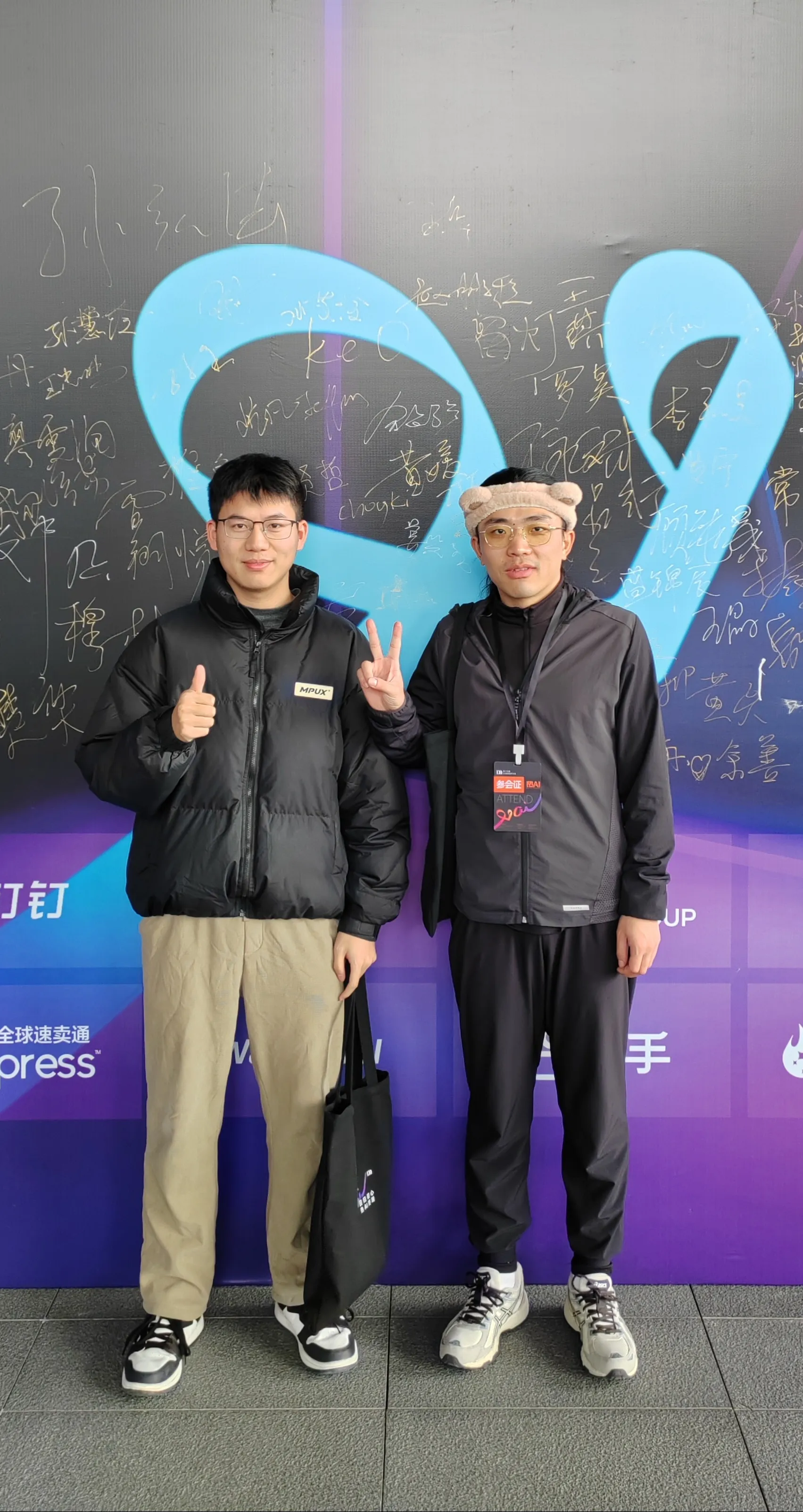 前面站着的是作者本人和师兄，两位站在大会签名背景板前的人，左边的人竖大拇指，右边的人比出剪刀手；背景板上布满参会者手写签名，并有蓝色艺术图案装饰，传递会议互动氛围。