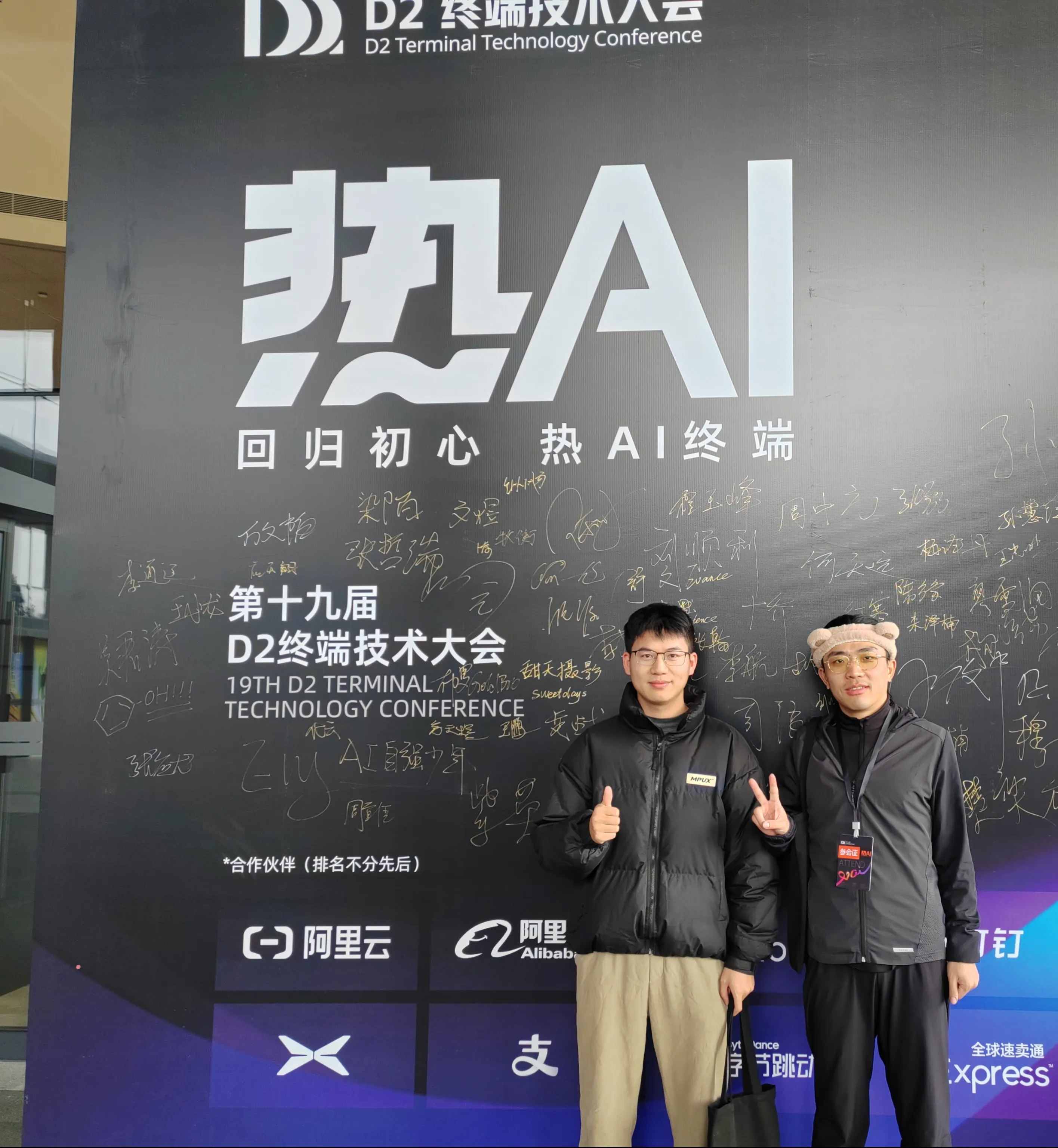 两人站在 “第十九届 D2 终端技术大会” 背景板前，背景板显著位置写着 “热 AI 回归初心 热 AI 终端”，下方标注会议名称，底部展示阿里云、支付宝等合作伙伴标识，记录会议留念瞬间。