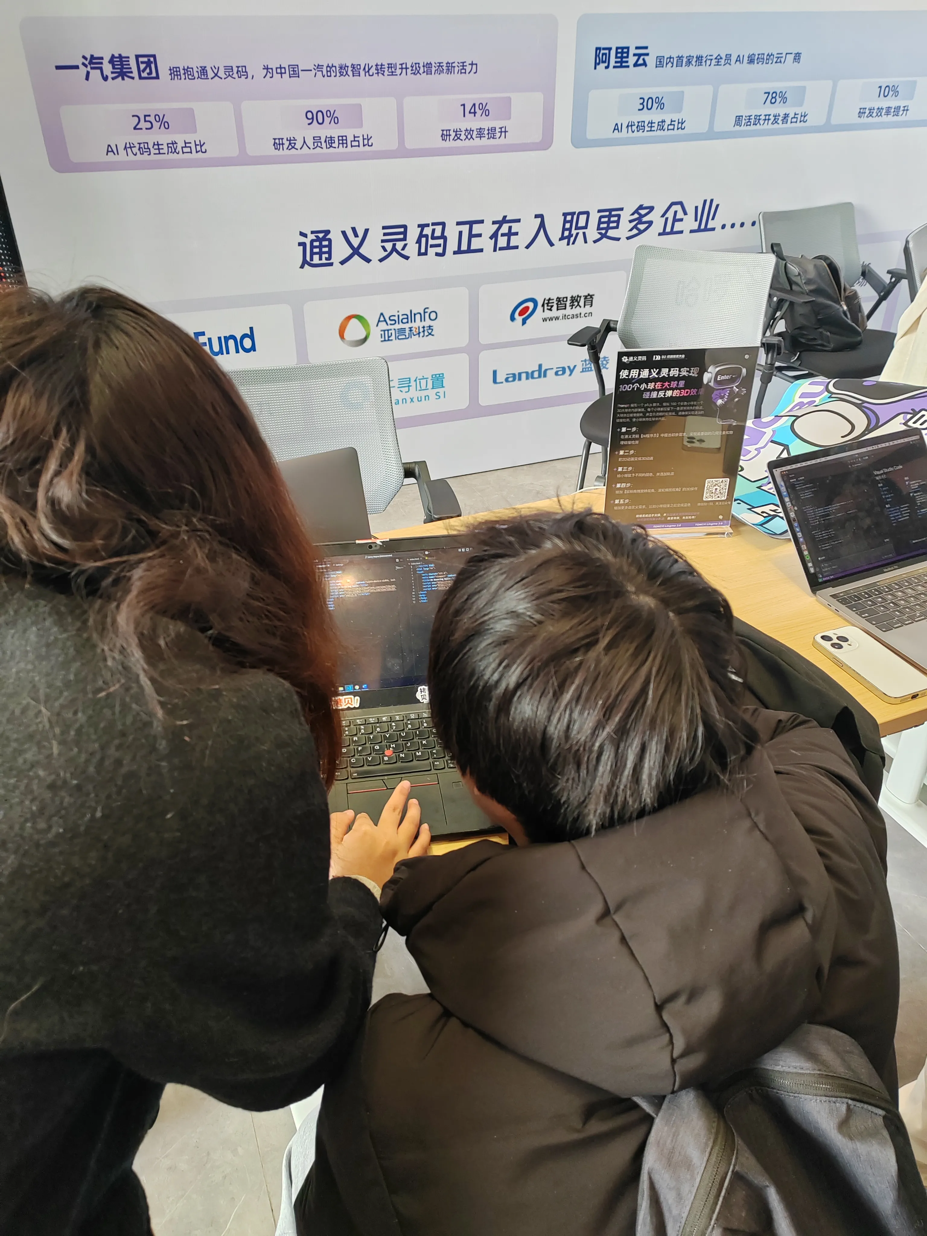 展会现场，两位参会者正俯身查看笔记本电脑内容，背景展板展示 “通义灵码” 合作企业数据，如 “一汽集团 AI 代码生成占比 25%”“阿里云 AI 代码生成占比 30%” 等信息，呈现技术交流场景。
