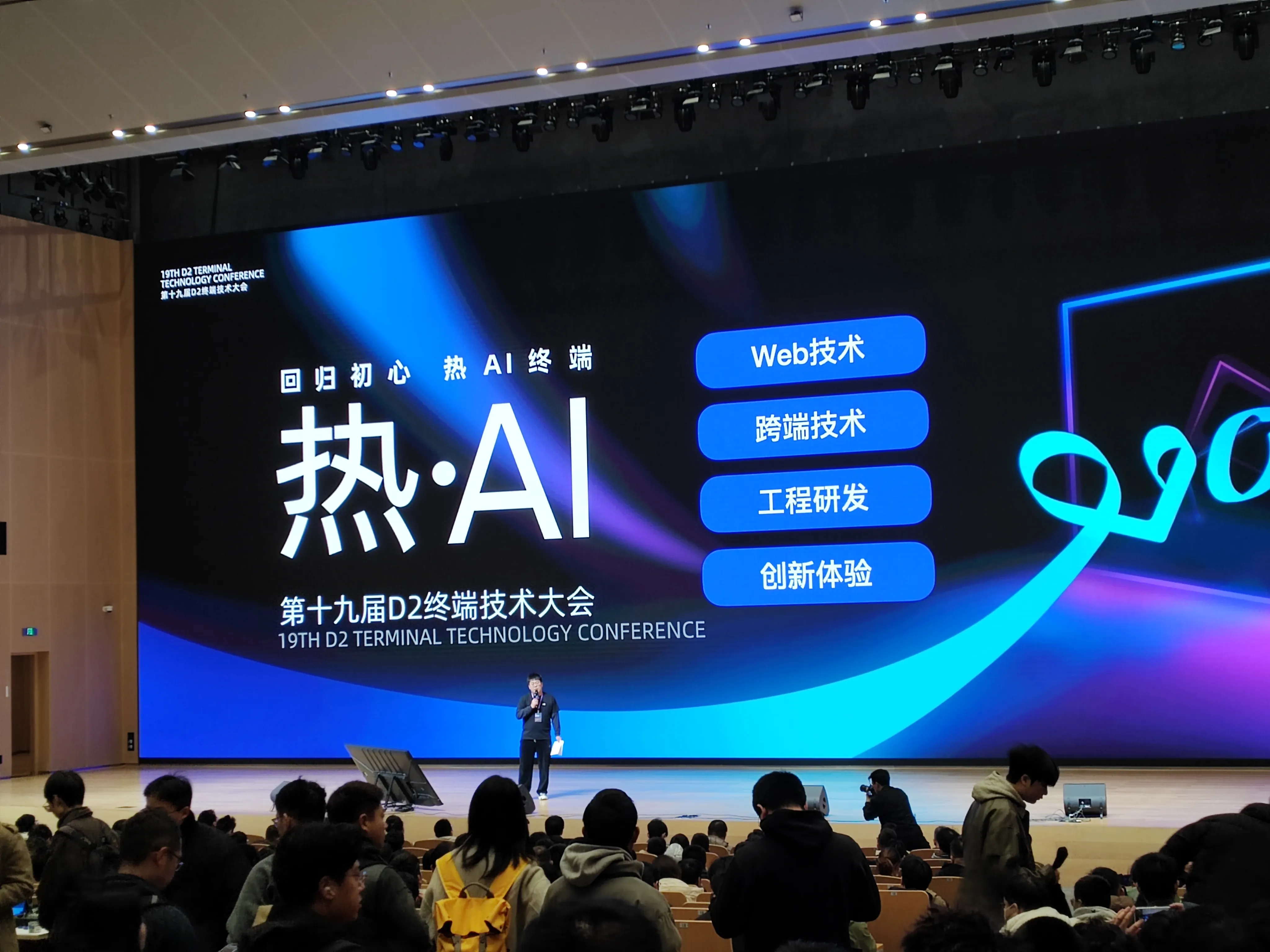 第十九届 D2 终端技术大会现场，巨大的舞台背景屏显示 “回归初心 热 AI 终端”“热・AI” 主题，以及 “Web 技术”“跨端技术”“工程研发”“创新体验” 分论坛板块，一位演讲者站在舞台中央，台下坐满参会观众，顶部灯光设备清晰可见，呈现专业会议氛围。