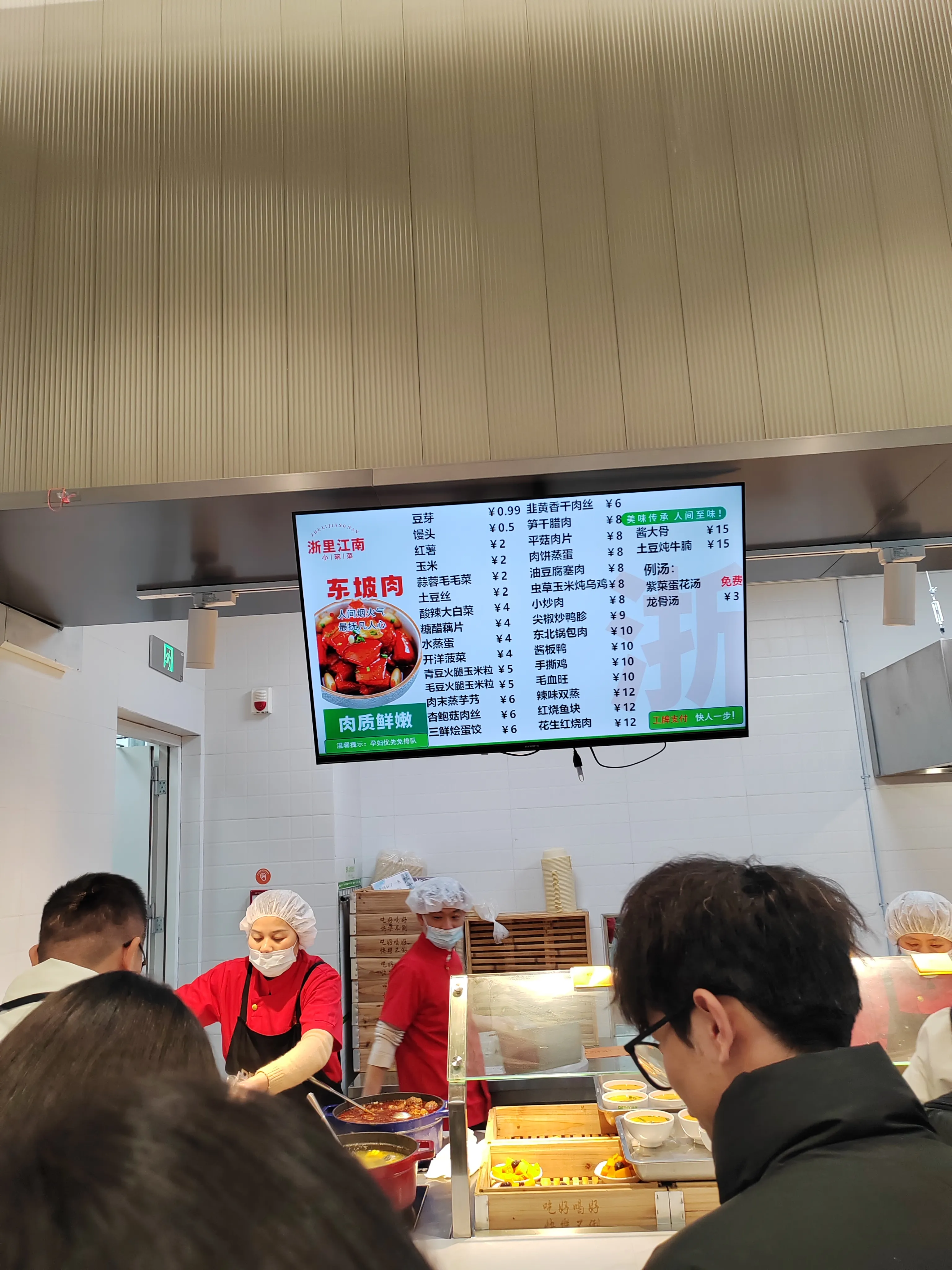 阿里巴巴餐厅物价表，食堂环境中，上方电子屏显示 “浙里江南 东坡肉” 菜单，罗列豆芽、银头、玉米等菜品及价格（如豆芽 0.99 元、玉米 2 元），工作人员穿着红色制服、戴着卫生帽操作餐食，顾客在窗口排队等待点餐。