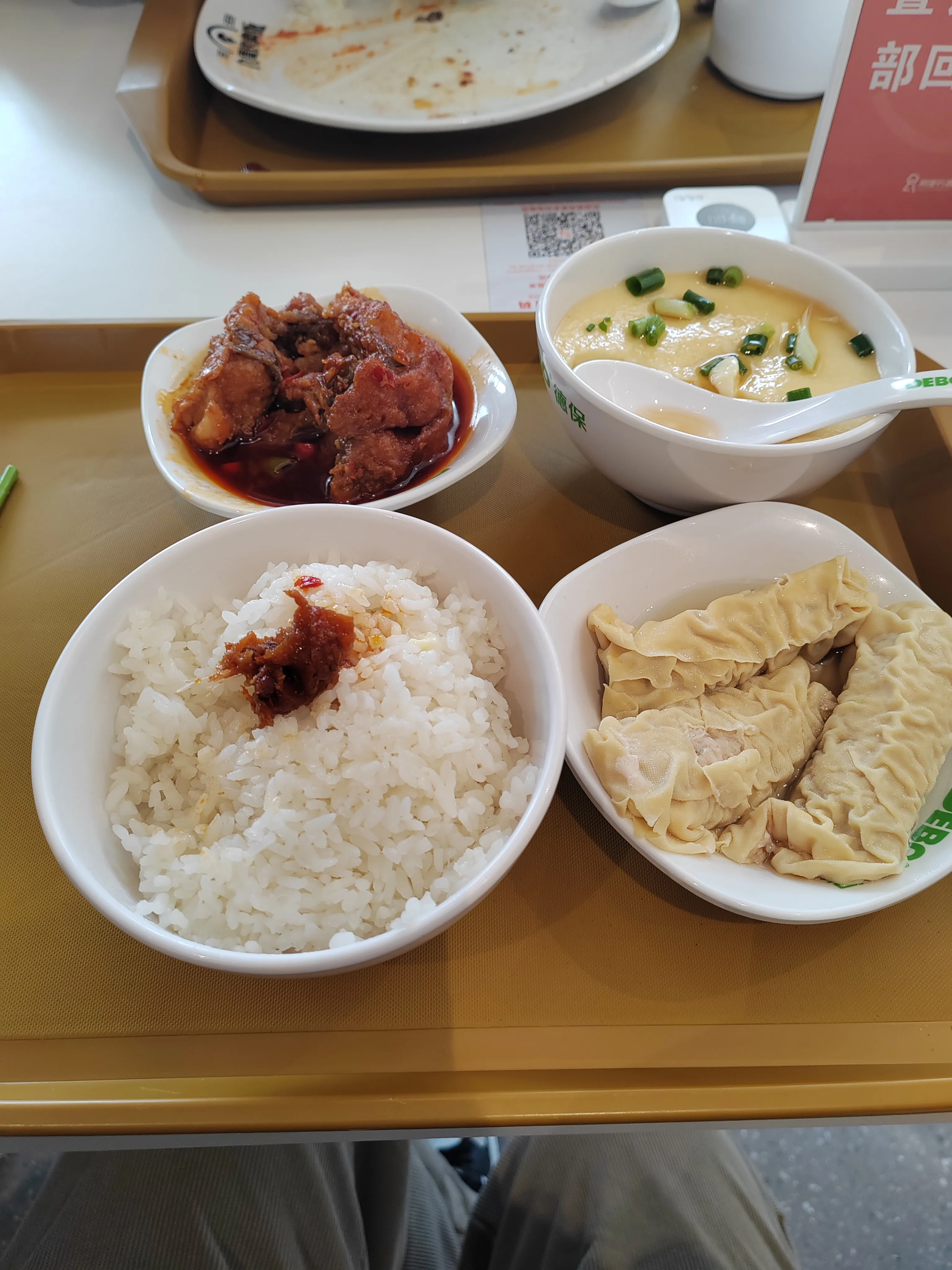 托盘上摆放着一份餐食，包括白米饭（顶部点缀少量鱼肉）、红烧鱼肉、蒸蛋（撒有葱花）、豆皮包裹的肉卷，呈现食堂用餐的具体食物搭配。