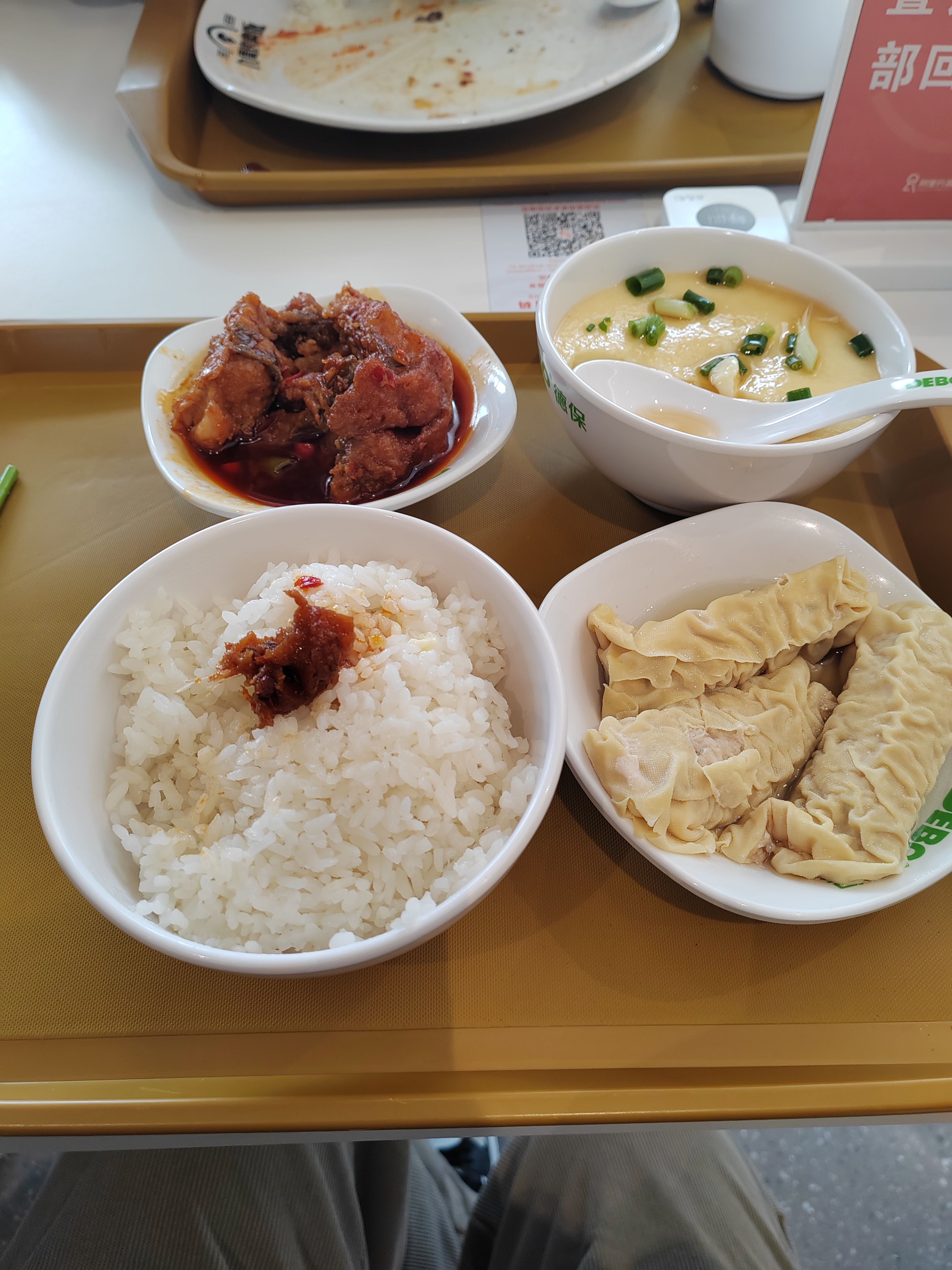 托盘上摆放着一份餐食,包括白米饭(顶部点缀少量鱼肉)、红烧鱼肉、蒸蛋(撒有葱花)、豆皮包裹的肉卷,呈现食堂用餐的具体食物搭配。
