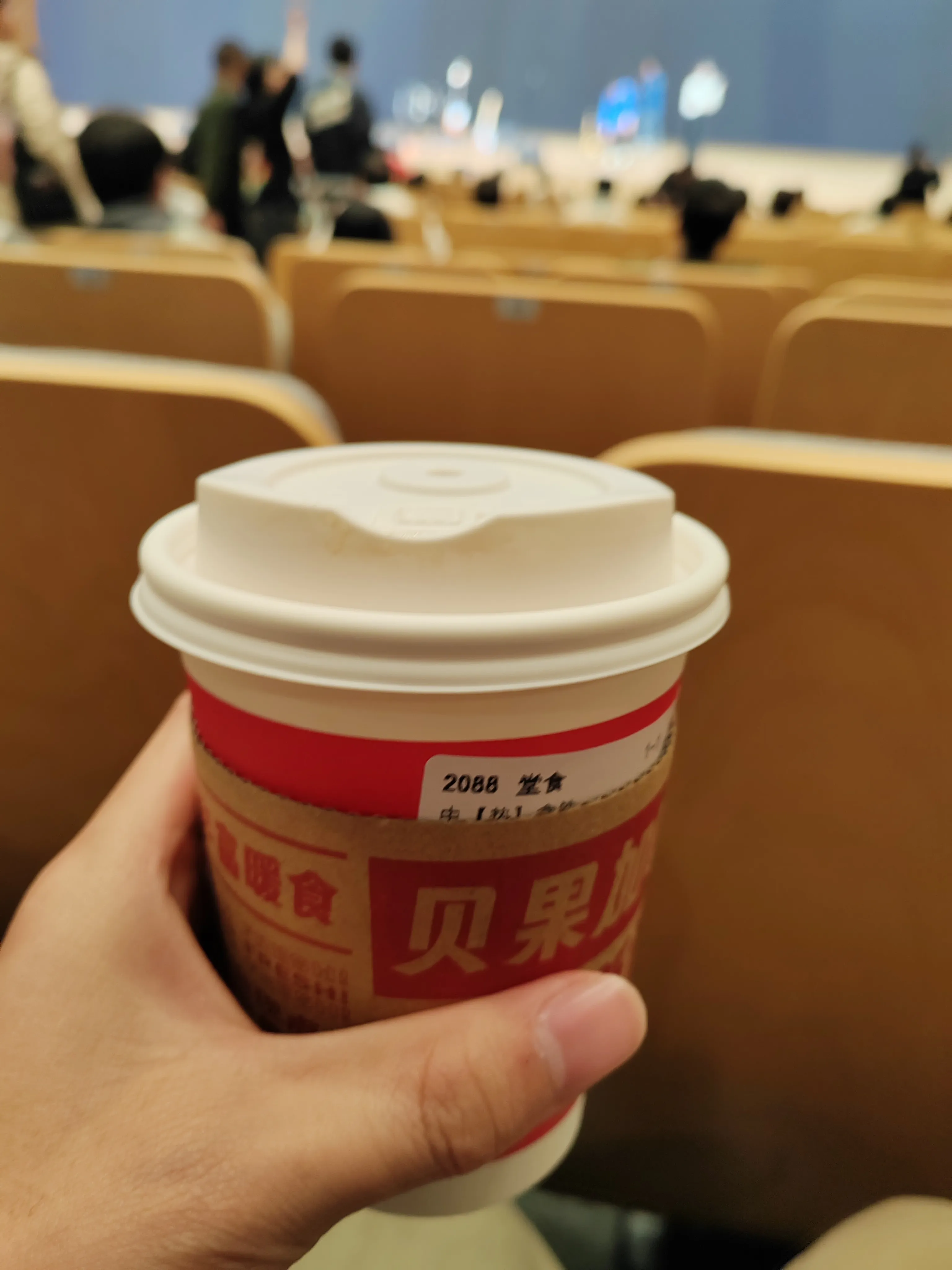 一杯拿铁，一只手握着带白色杯盖的饮品杯，杯身标签有红色文字，背景模糊显示会议现场的棕色座椅，暗示在会议场所内手持饮品的情景。