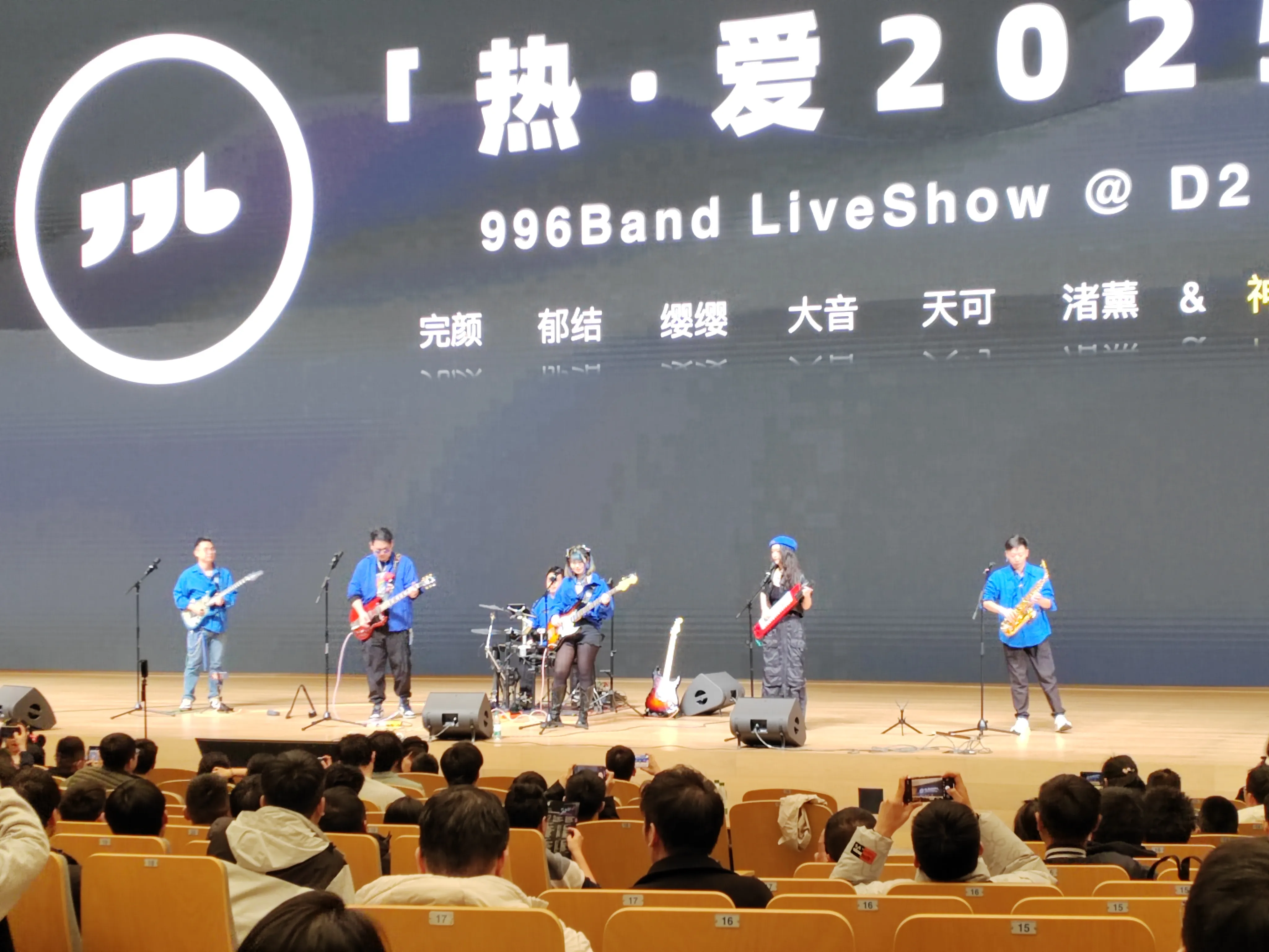 996乐队，舞台上 “996Band LiveShow @ D2” 演出，背景屏显示 “热・爱 2025”，乐队成员穿着蓝色服装，分别演奏吉他、架子鼓、萨克斯等乐器，台下观众举起手机拍摄，氛围热烈。