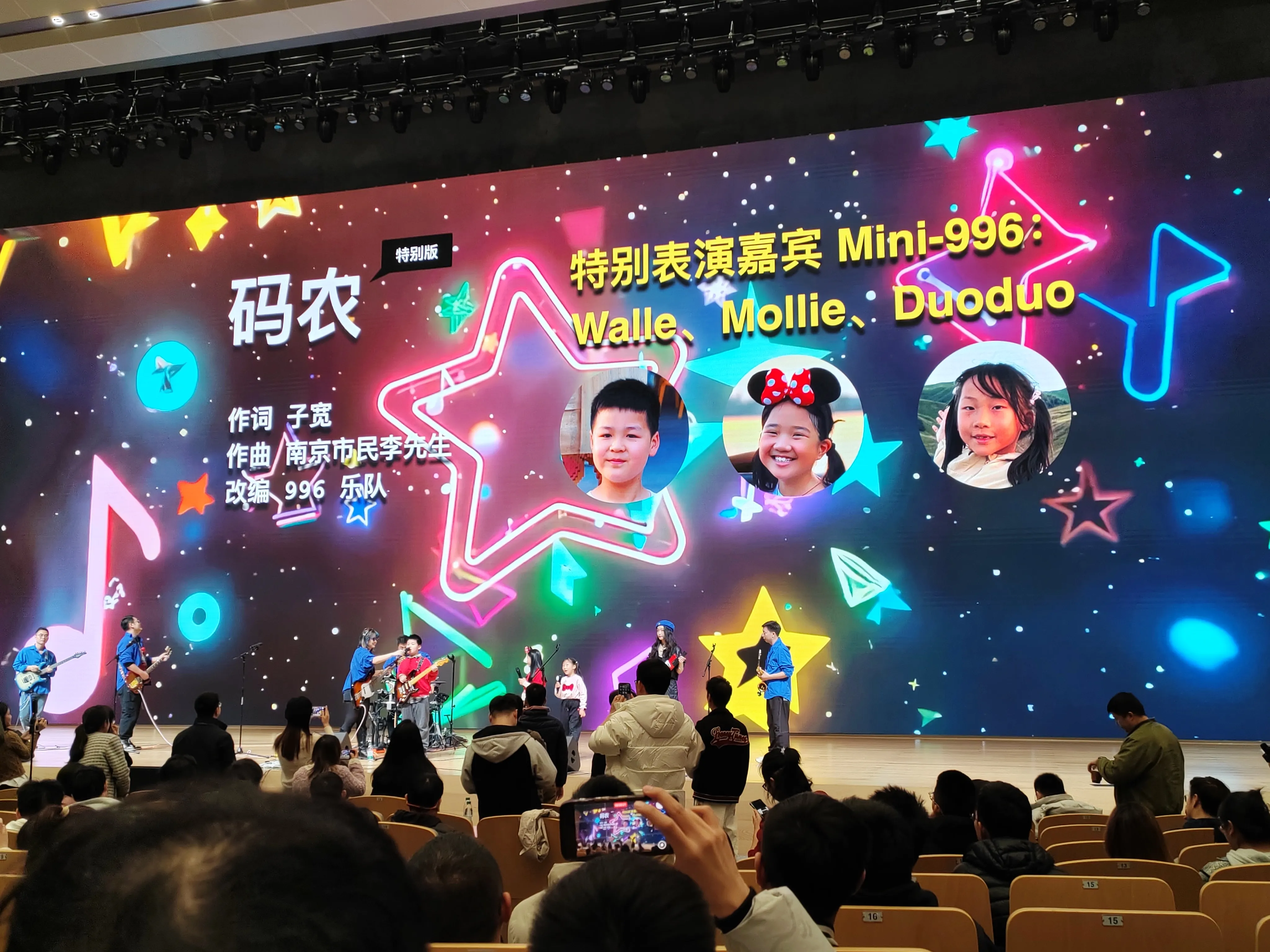 mini-996乐队，舞台背景色彩绚丽，有星星、音符装饰，标注 “特别表演嘉宾 Mini-996：Walle、Mollie、Duoduo” 及嘉宾照片，乐队与小朋友共同表演，台下观众聚焦舞台，部分人拍摄记录