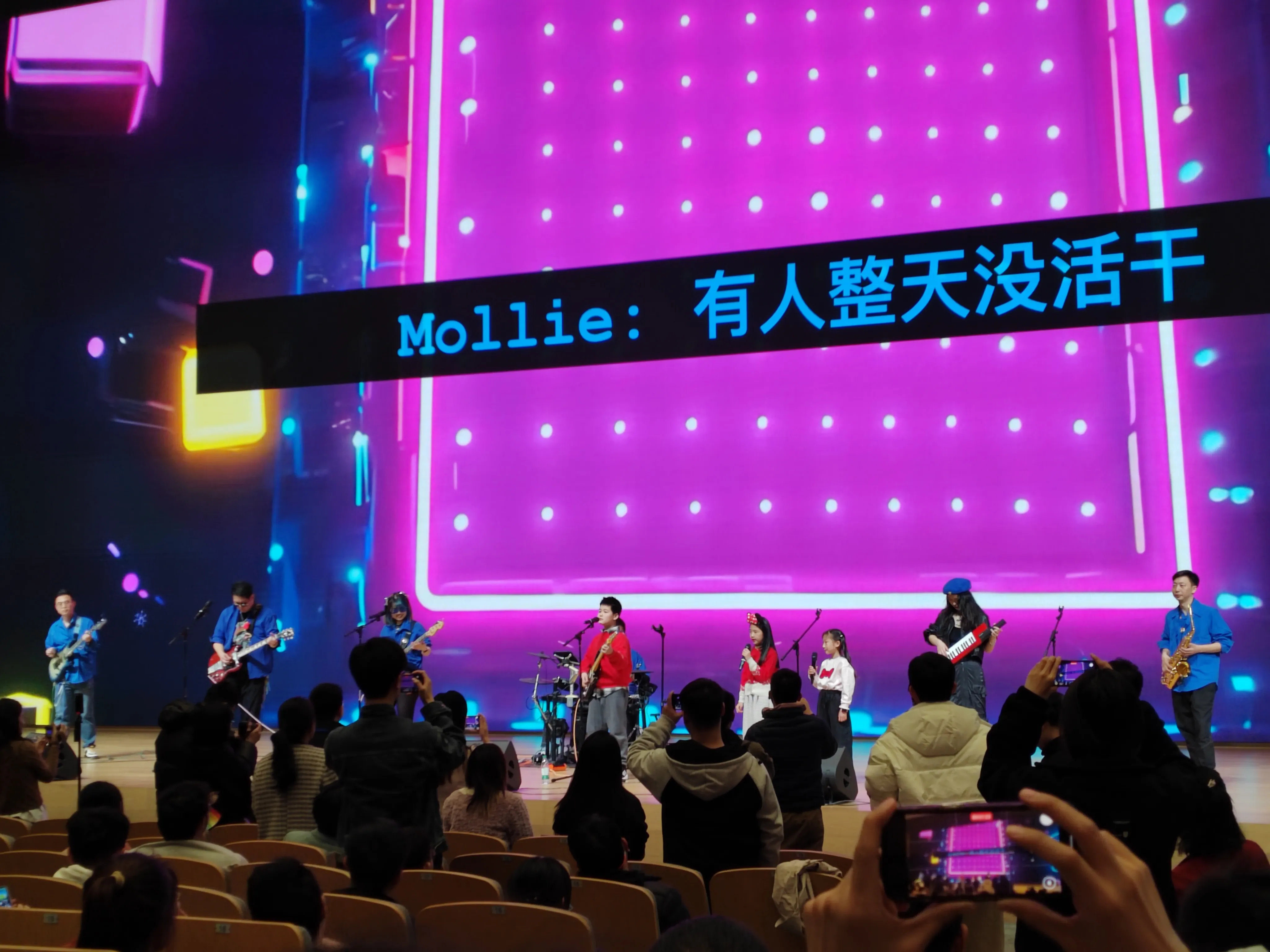 mini-996乐队,舞台背景为紫色灯光特效，上方字幕显示 “Mollie：有人整天没活干”，乐队成员继续演奏，观众坐在棕色座椅上观看，部分人举起手机录制演出画面，展现互动性演出环节。