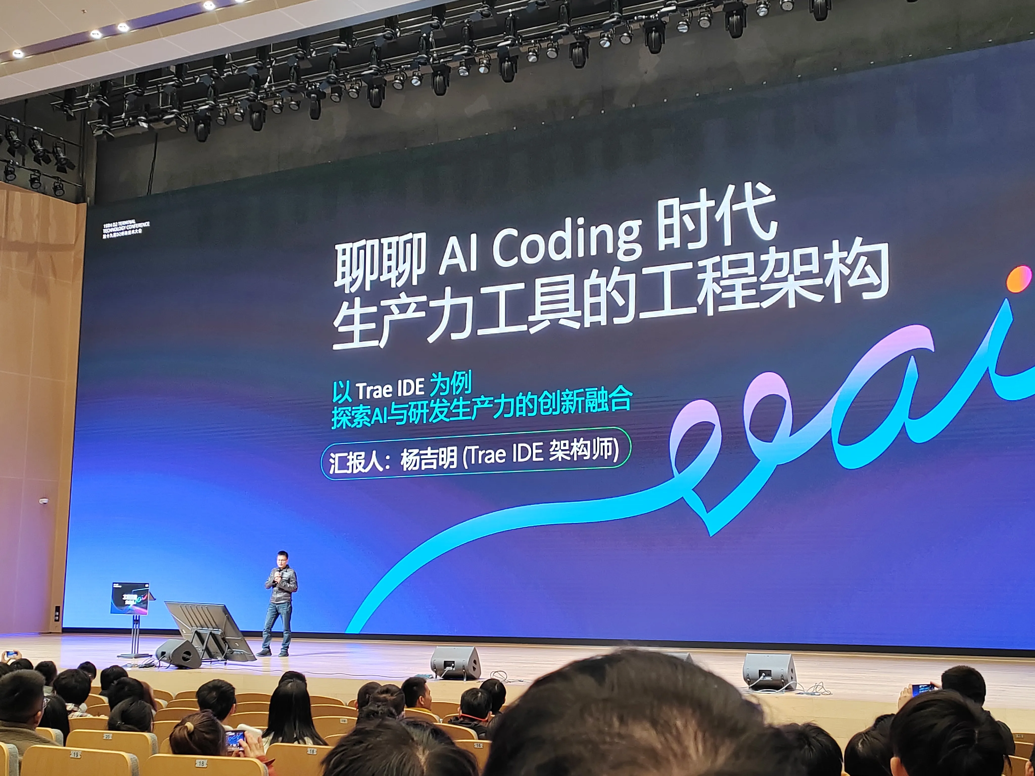 会议现场，巨大背景屏显示 “聊聊 AI Coding 时代生产力工具的工程架构”，副标题标注 “以 Trae IDE 为例 探索 AI 与研发生产力的创新融合”，汇报人 “杨吉明（Trae IDE 架构师）” 站在舞台中央讲解，台下坐满专注聆听的参会观众，顶部灯光设备清晰可见。