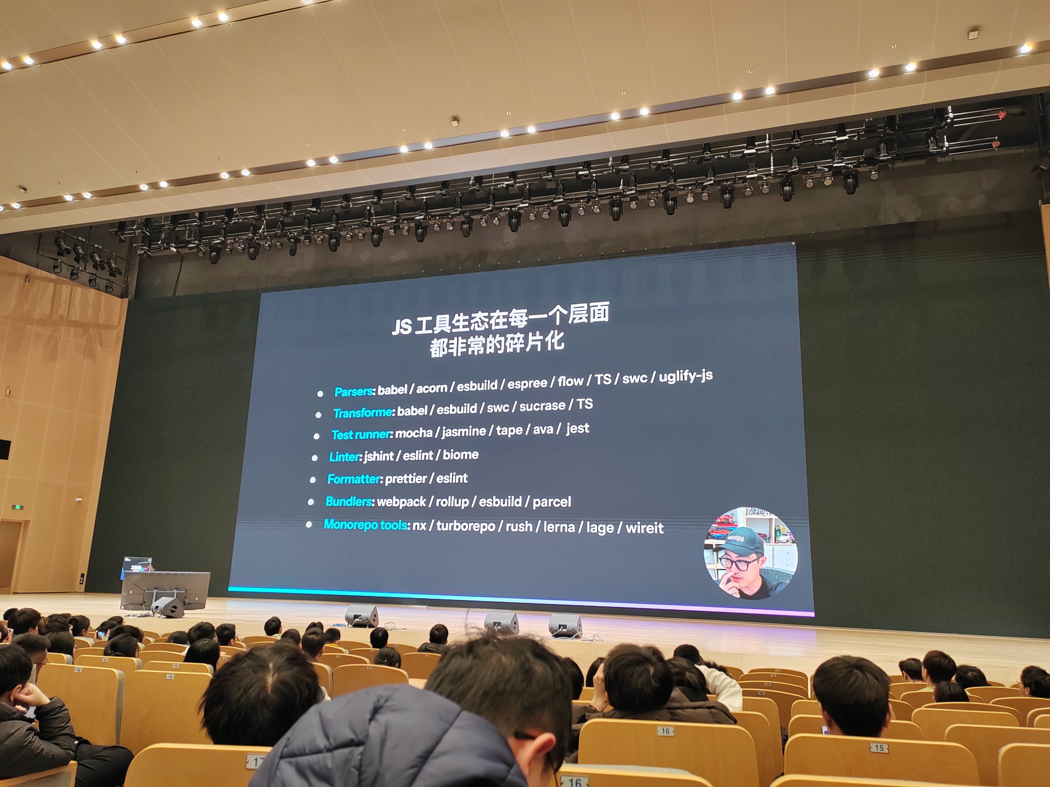 vuejs作者尤雨溪聊前端工具链的问题,背景屏标题 “JS 工具生态在每一个层面都非常的碎片化”,详细罗列解析器(babel、esbuild 等)、转换器、测试工具等不同类别的工具列表,观众坐在会议座椅上专注听讲。