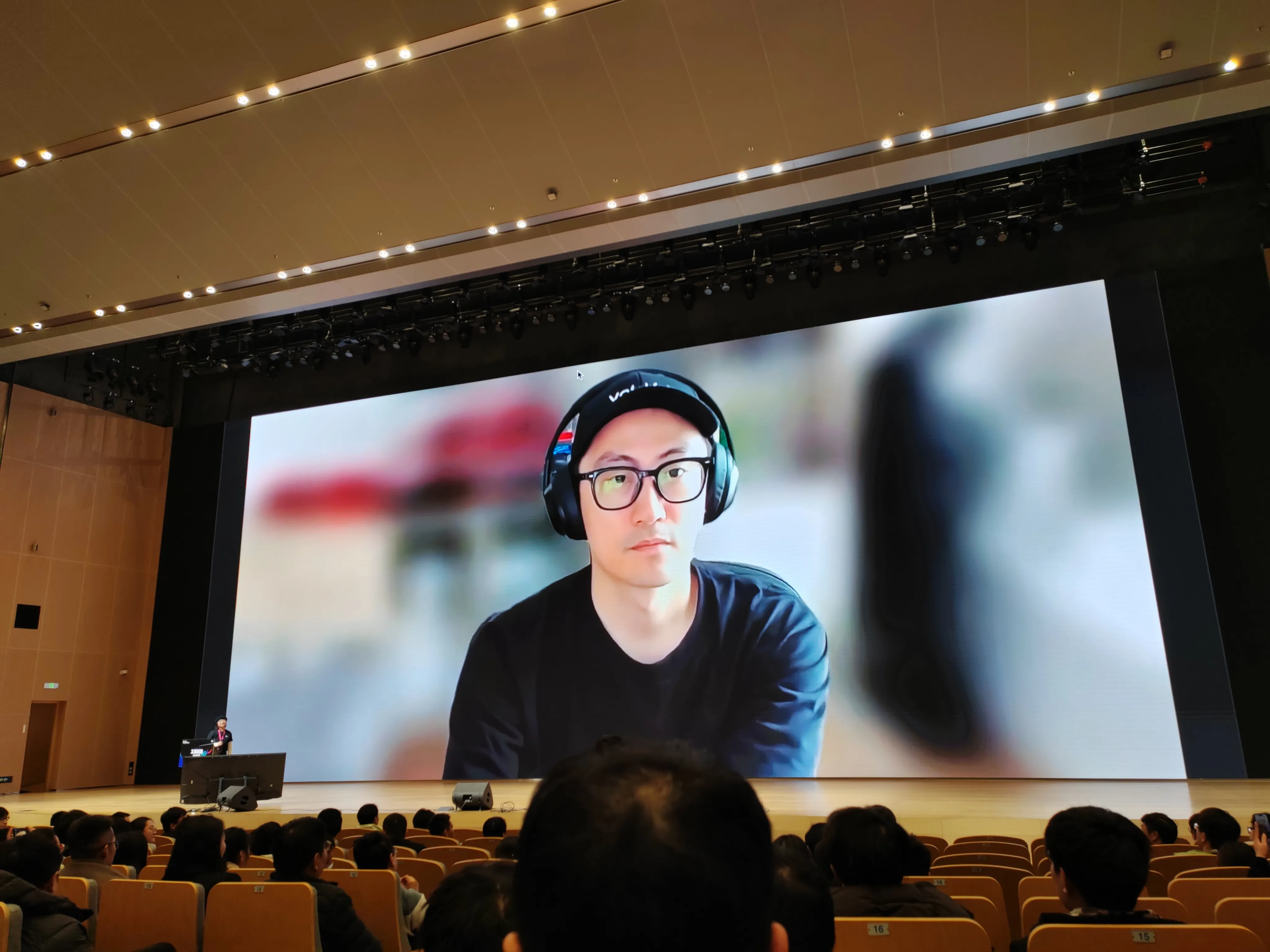 尤雨溪答观众提问,会议舞台背景屏显示一张人物照片，人物戴着耳机，背景模糊处理，台下观众处于会议场景中，可能为演讲关联的人物介绍环节。