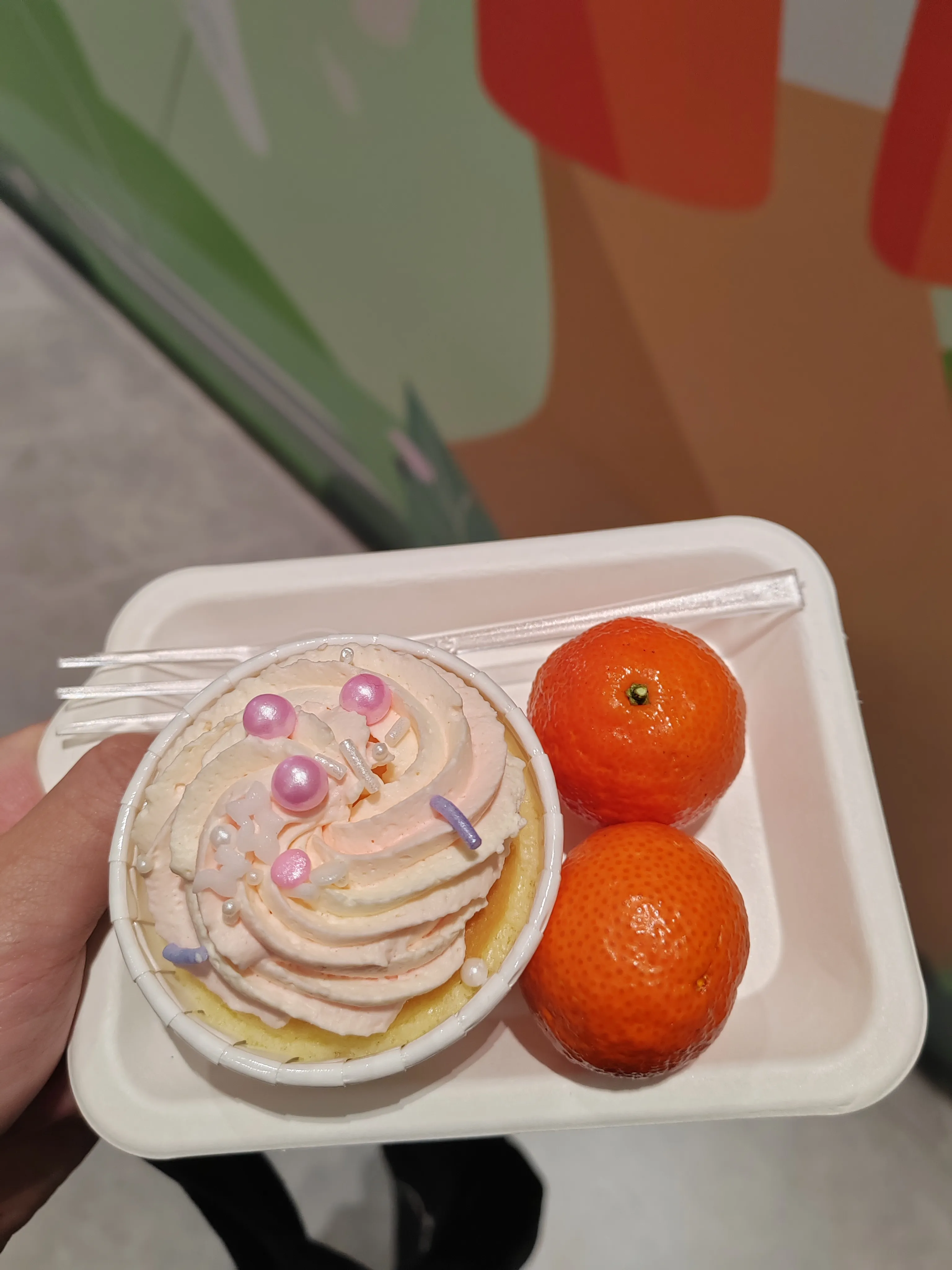 一只手托着白色托盘，托盘上有一份装饰粉色奶油和珍珠糖的甜点，旁边摆放两个橙色橘子，配透明餐具，呈现会议期间的茶歇食品。