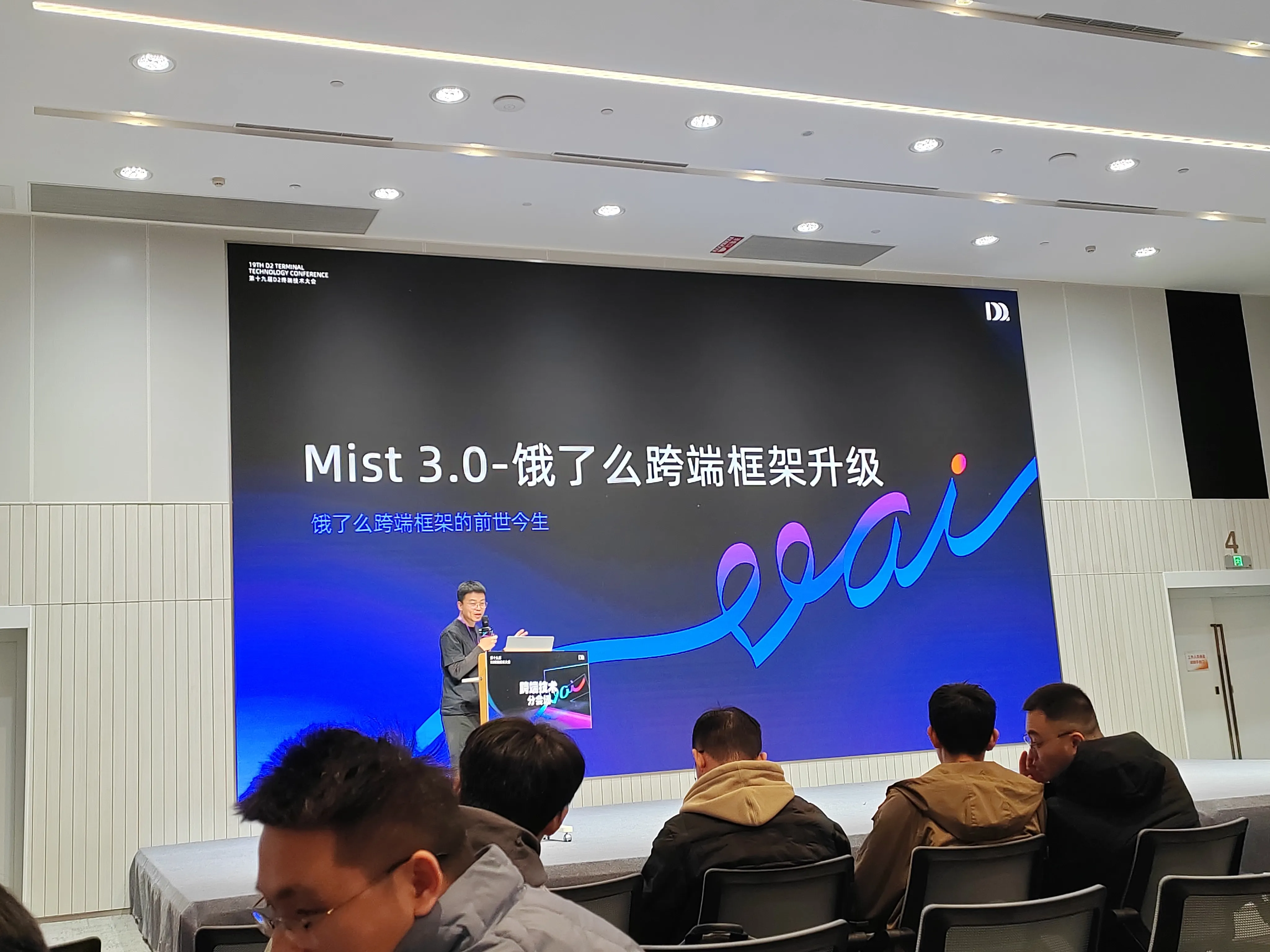 饿了么主创团队讲解mist3.0框架,分会场场景，背景屏显示 “Mist 3.0 - 饿了么跨端框架升级”，副标题 “饿了么跨端框架的前世今生”，演讲者站在讲台后讲解，几位参会者坐在前排聆听。