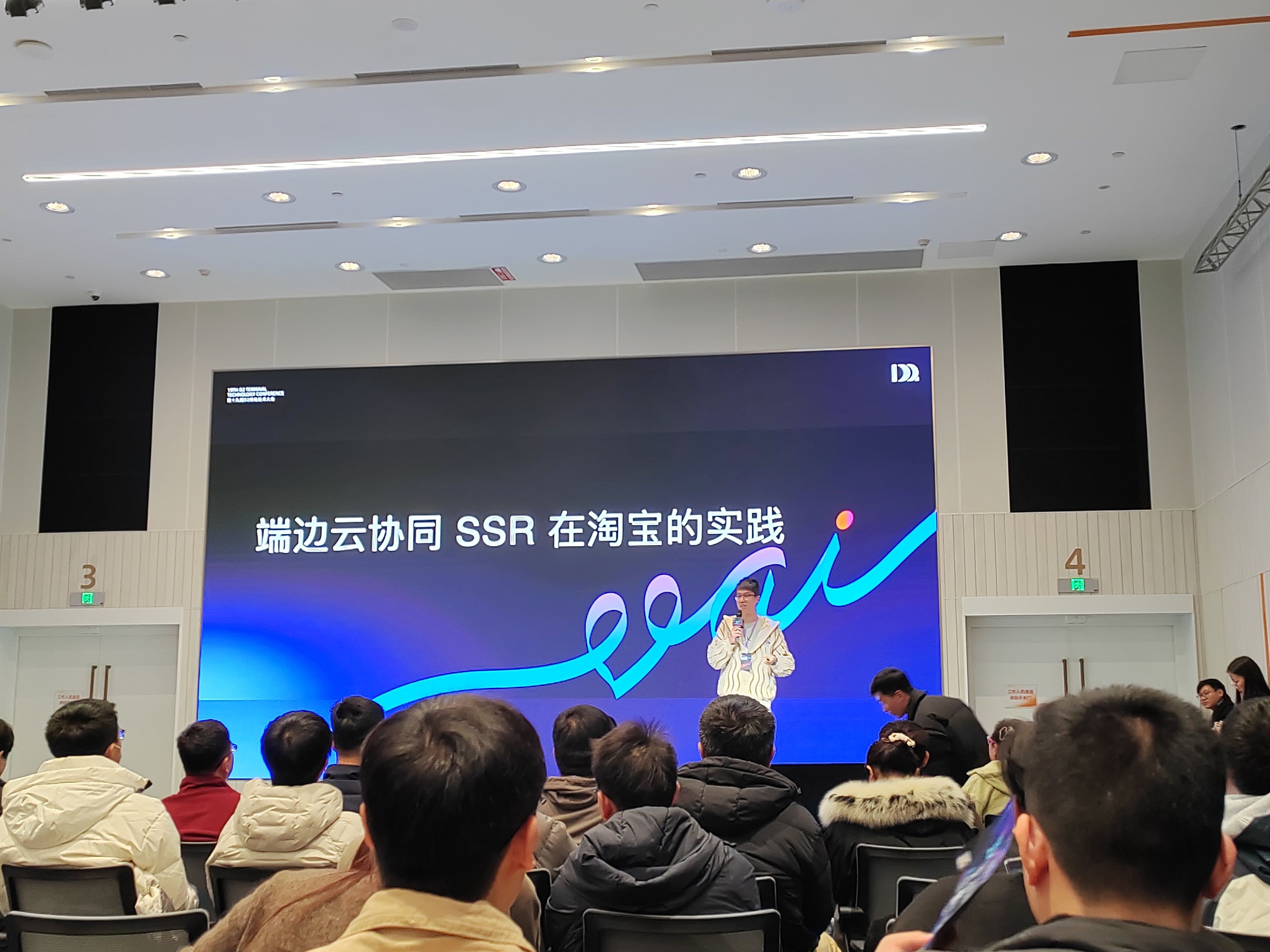 演讲主题为 “端边云协同 SSR 在淘宝的实践”,背景屏以蓝紫色渐变为主,搭配会议主题装饰图案。演讲者站在舞台中央讲解,台下参会者密集就座,部分人手持资料,现场氛围专注,聚焦淘宝在端边云协同 SSR 领域的实践分享。