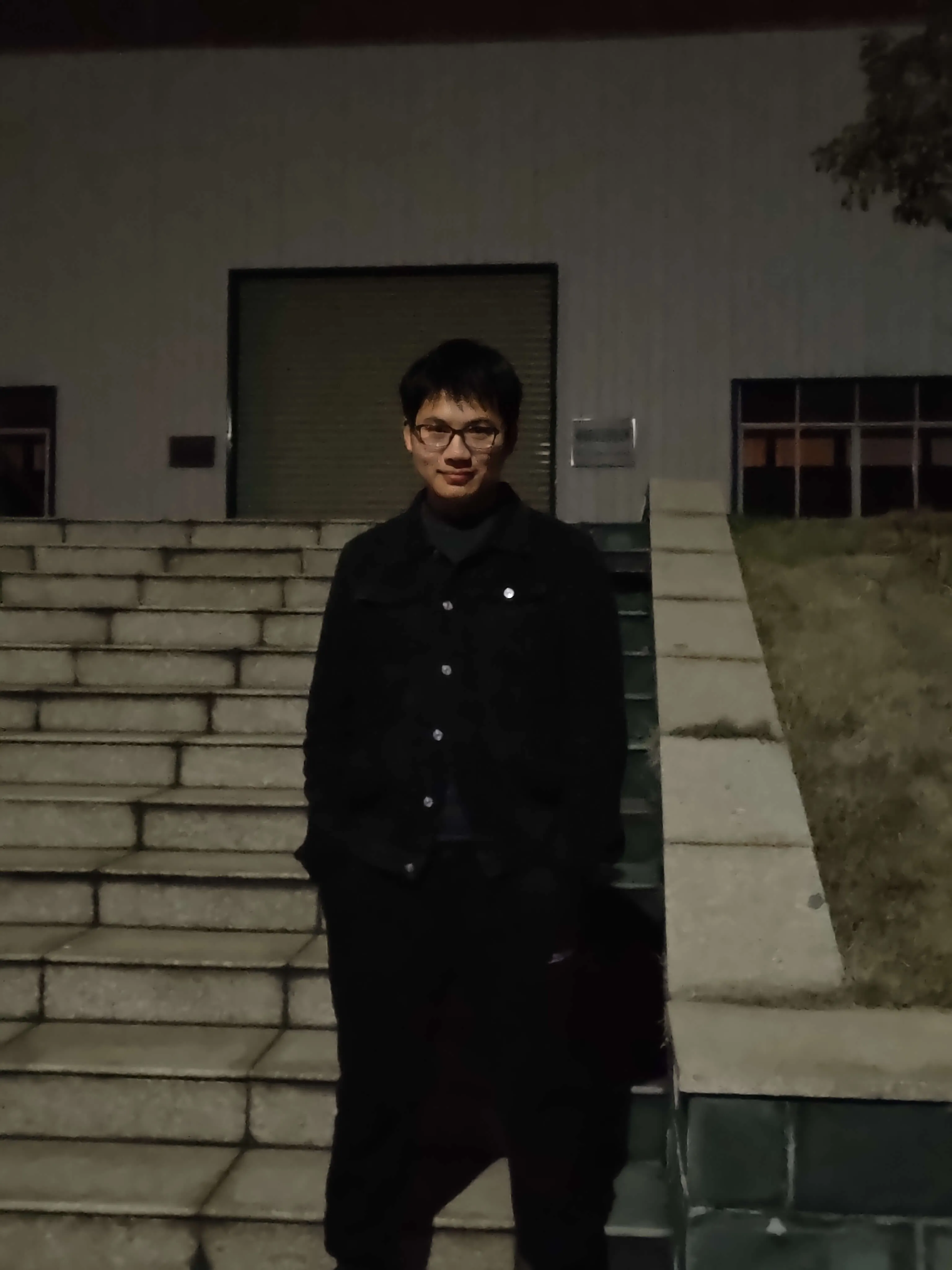 2023-2-18日新余学院拍摄。夜晚场景中,人物身穿黑色外套与黑色长裤,双手插兜站在台阶前。台阶由浅灰色石块铺成,后方是浅色建筑墙体,墙体有深色卷帘门和亮灯的窗户,右侧可见草地斜坡,氛围静谧。