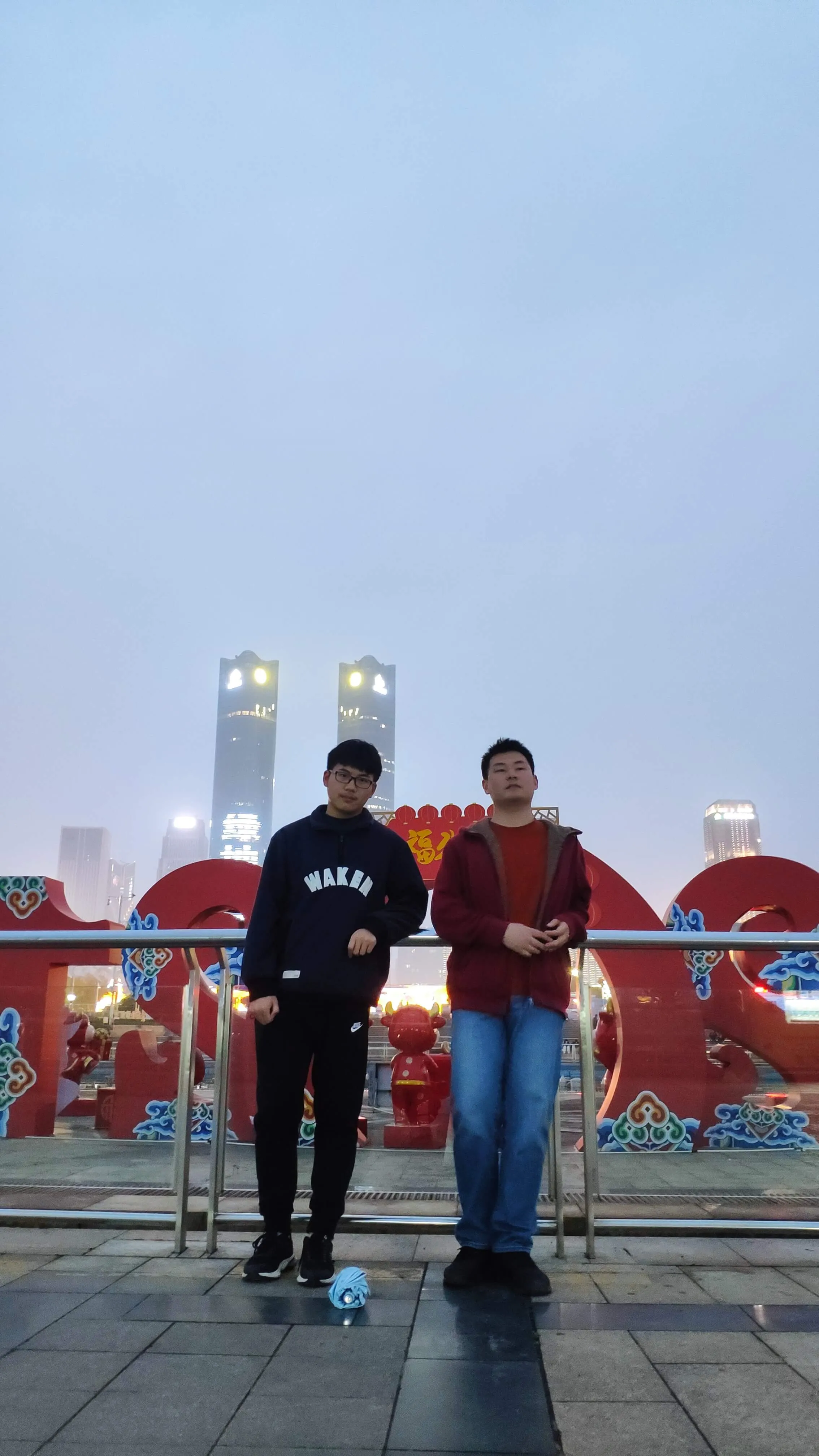 2021-3-21日与好友张合照 人物:两位男性站立在画面中央,左侧的男性穿着深色连帽衫和黑色裤子,戴着眼镜;右侧的男性穿着红色外套和蓝色牛仔裤。
背景:背景中有一个大型的红色装饰物,形状类似于汉字“福”,装饰物上有蓝色的花纹图案。远处可以看到几座高楼大厦,其中两座高楼上亮着灯光,天空呈现出淡淡的蓝色,显得有些阴沉。
地面:他们站立在一个铺有灰色地砖的平台上,地面上有一把浅蓝色的雨伞。在一个城市广场