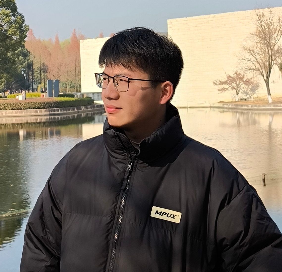 作者2025年1月在杭州良渚博物馆前湖边拍摄，一位站在户外的男性。他穿着一件黑色的羽绒服，羽绒服上有一个标有“MPUX”字样的标签。他戴着眼镜，头发短而整齐。背景中可以看到一个平静的水面，水面上反射着周围的景色。远处有一些建筑物和树木，树木的颜色呈现出秋天或冬天的色调，树叶稀疏，部分树枝裸露。整体环境显得宁静而开阔，天空晴朗，光线柔和。